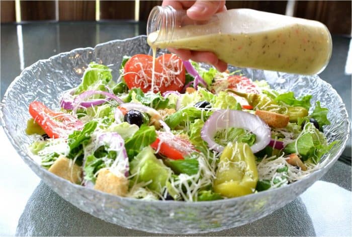 This homemade copycat Olive Garden Salad Dressing tastes just like the restaurant version. Learn how to make this salad at home for your family and friends! #copycatrecipe #restaurantcopycats #italianfood #italiansidedishes #sidedishideas #easyrecipes copycat olive garden salad dressing being poured over a large glass bowl filled with olive garden salad.