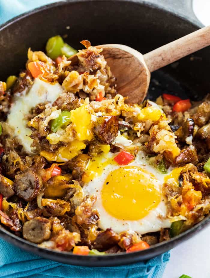 This breakfast skillet is loaded with crispy golden hash, mouth-watering sausage, and perfectly cooked eggs. All you need is a single skillet to make this deliciously satisfying breakfast! | The Cozy Cook | #eggs #hash A cast iron skillet with a baked egg and a wooden spoon stirring cooked hash, sausage, and bell peppers.