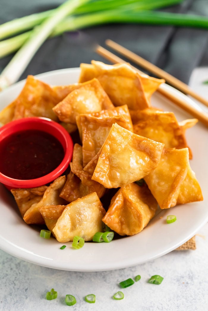 Crispy wontons are baked or fried to golden perfection and filled with an easy cream cheese Crab Rangoon filling. Plus, they're easy to make ahead of time for seamless entertaining! A white plate filled with fried crab rangoon and a red ramekin of sweet and sour sauce with chopsticks and green onions in background.