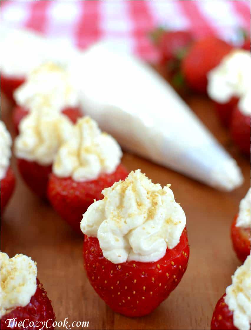 These fresh and flavorful strawberries are stuffed with an easy, creamy cheesecake filling! This is an elegant and fun way to serve dessert for special occasions with friends and family! | The Cozy Cook | #cheesecake #strawberries Strawberries standing on a wooden board and stuffing with cheesecake filling with white piping bag in background.