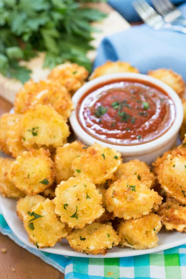 Cheese-filled tortellini that’s surrounded by a crispy Parmesan crust, fried to golden perfection and dipped in warm marinara sauce. a plate of breaded, fried tortellini with marinara sauce in background.