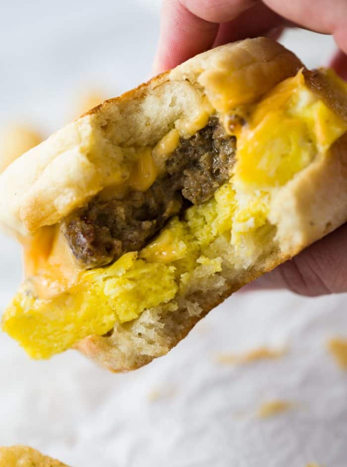 Crisp and buttery English muffins topped with fluffy eggs, bacon, sausage, and cheese. Easy to freeze ahead of time and heat in just 3 minutes! A close up view of a hand holding a sausage egg and cheese sandwich with a bite taken out of it.
