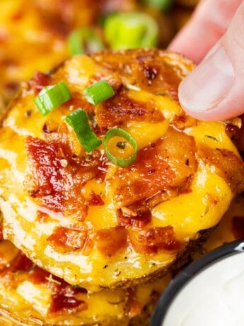 A hand holding a crispy slice of potato topped with melted cheese, bacon, and green onions.
