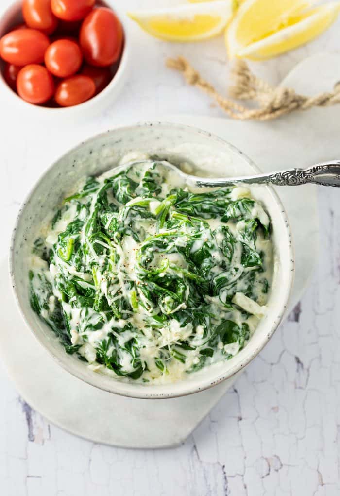 Swirls of fresh spinach combined in an indulgent Parmesan cream sauce. This is a perfect side dish on its own or as a topping on chicken and fish. It even makes a delicious addition to casseroles! A white bowl filled with creamed spinach on a white table next to cherry tomatoes and sliced lemons.