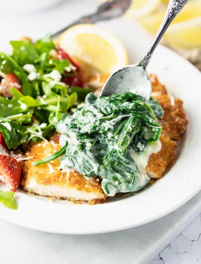 Swirls of fresh spinach combined in an indulgent Parmesan cream sauce. This is a perfect side dish on its own or as a topping on chicken and fish. It even makes a delicious addition to casseroles! A spoon spreading creamed spinach on top of breaded chicken