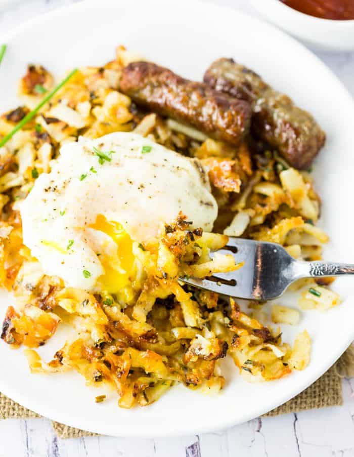 Extra crispy, perfectly golden hash browns combined with onions, cheese, and chives and cooked in just minutes right in your waffle iron! A fork scooping up waffle iron hash browns with an egg on top on a white plate.
