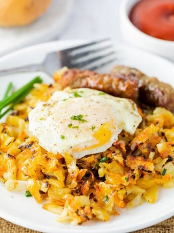 A white plate with waffle iron hash browns topped with a fried egg with sausage links in the background.
