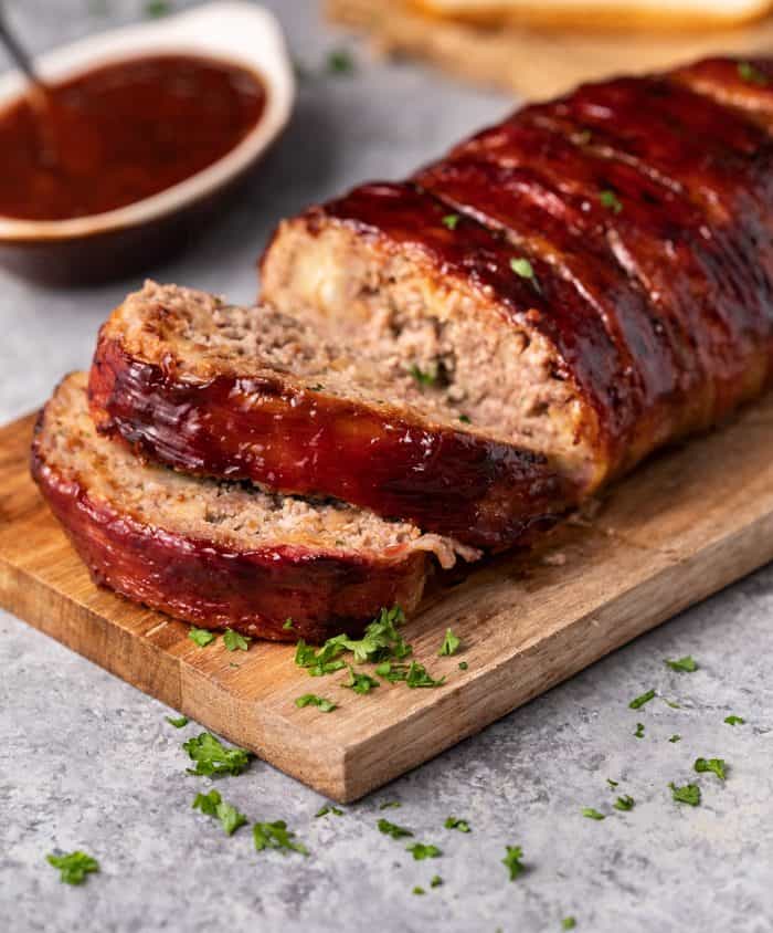 This Pioneer Woman Meatloaf Recipe is the best you'll ever try! The entire loaf is wrapped in bacon and baked to perfection, and it freezes well for future meals! | The Cozy Cook | #Meatloaf #ThePioneerWoman #Dinner #Mealprep #Beef Meatloaf sliced on a wooden cutting board with fresh parsley sprinkled on top and homemade ketchup sauce in background