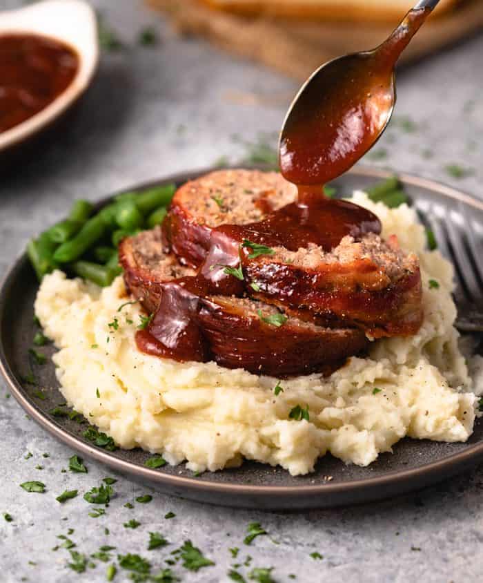 This Pioneer Woman Meatloaf Recipe is the best you'll ever try! The entire loaf is wrapped in bacon and baked to perfection, and it freezes well for future meals! | The Cozy Cook | #Meatloaf #ThePioneerWoman #Dinner #Mealprep #Beef Meatloaf over mashed potatoes with a homemade ketchup sauce drizzled on top
