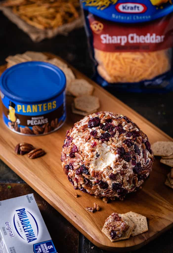 This sweet and savory Pineapple Pecan Cheese Ball has an unbeatable flavor combination that includes pineapple, pecans, bacon, two cheeses, and more! It’s ready in minutes, can be made ahead of time, and can even be baked! | The Cozy Cook | #Appetizer #Cheeseball Wooden Cutting Board with Pineapple Cranberry Cheese Ball with planters pecans and grated cheese in background.