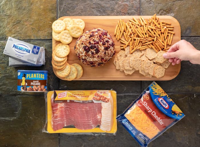 This sweet and savory Pineapple Pecan Cheese Ball has an unbeatable flavor combination that includes pineapple, pecans, bacon, two cheeses, and more! It’s ready in minutes, can be made ahead of time, and can even be baked! | The Cozy Cook | #Appetizer #Cheeseball Wooden cutting board with cheese ball, crackers, pretzels, surrounding by a package of bacon, cheese, pecans, and cream cheese
