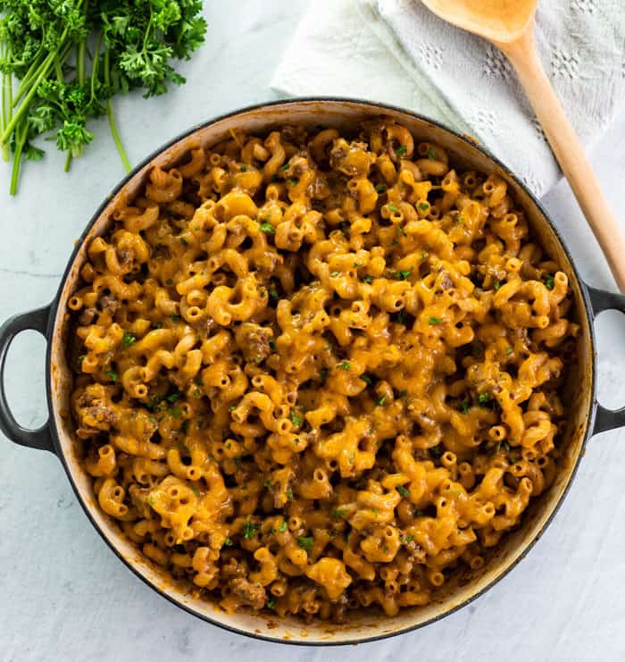 overhead view of a large skillet filled with warm cheeseburger casserole with a white background next to parsley and a wooden spoon.