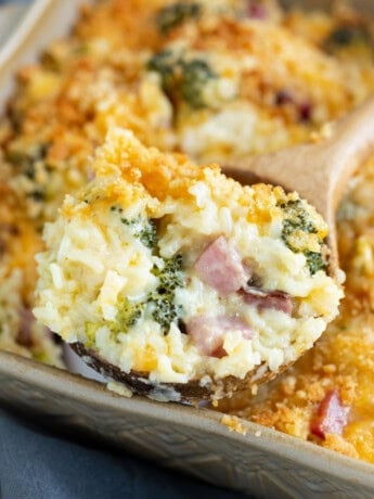 A wooden spoon scooping up ham and rice casserole with cheese from a casserole dish.