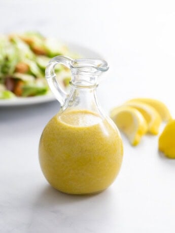 A glass bottle filled with homemade Caesar Dressing with a salad and lemon slices in the background.