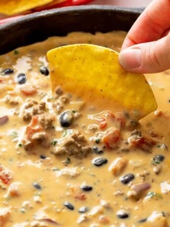 A hand dipping a tortilla chip into a skillet filled with Queso Dip with black beans, beef, and tomatoes.