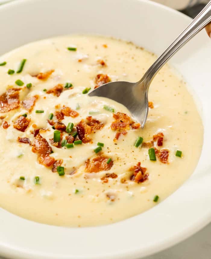 A white bowl with a spoon scooping up baked potato soup topped with bacon and chives.