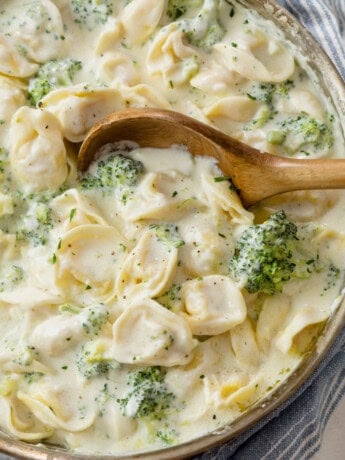 A wooden spoon in a skillet filled with Tortellini Alfredo with broccoli.