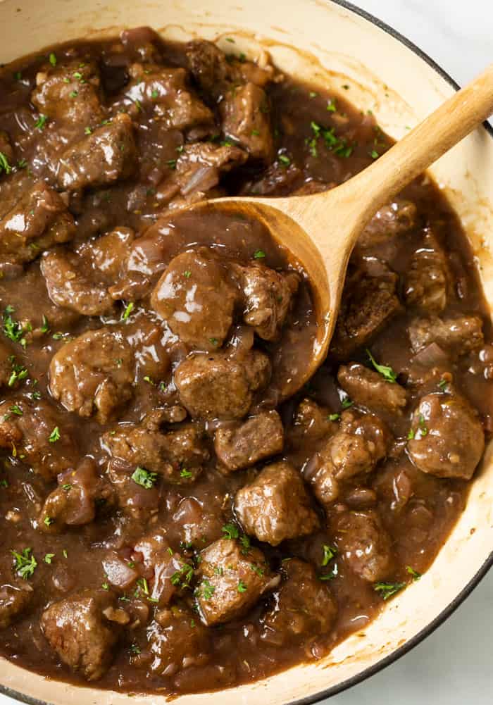 A wooden spoon scooping up Beef tips in gravy in a skillet topped with fresh parsley.
