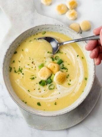 A white bowl with creamy potato leek soup in it with oyster crackers, green onions, and parsley on top.