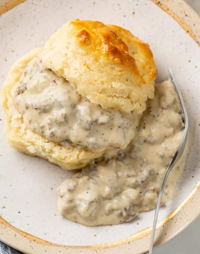 A buttermilk biscuit slice in half and topped with Sausage Gravy with a fork on the side.