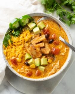 Overhead view of a white bowl with creamy chicken tortilla soup and garnishes like crispy tortillas, avocado, and shredded cheese.