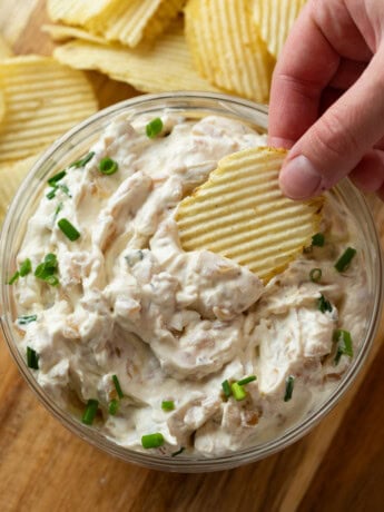 A hand dipping a Ruffled chip into a glass bowl of French Onion Dip topped with chives.