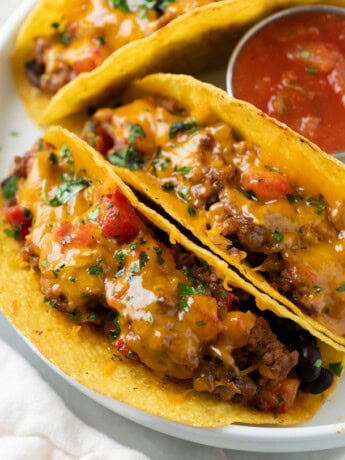 A white plate with baked tacos filled with ground beef and cheese with tomatoes.