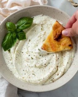 A bowl full of Feta Dip with a hand dipping bread into it with a garnish of basil.
