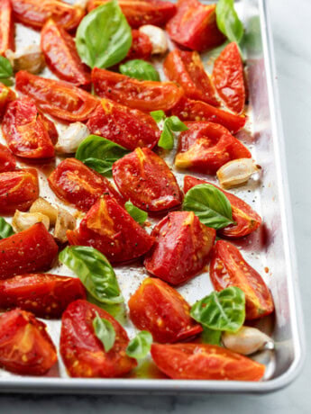 A rimmed baking sheet with roasted tomatoes and basil on top with salt and freshly cracked pepper.