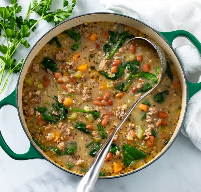 A green Dutch oven filled with Pinto Bean Soup with a soup ladle in it and fresh parsley on the side.