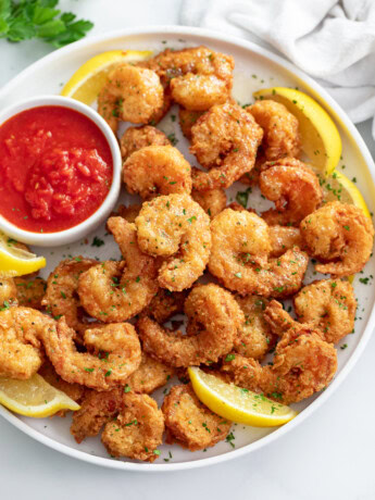 A pile of Fried Shrimp on a white plate with lemon slices and sauce.