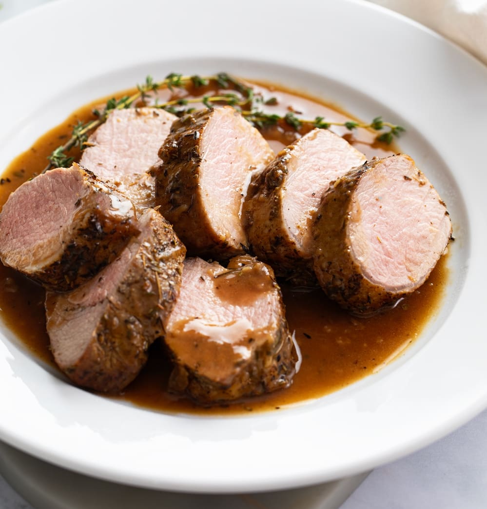Slices of Roasted Pork Tenderloin on a white plate with pan sauce and thyme.