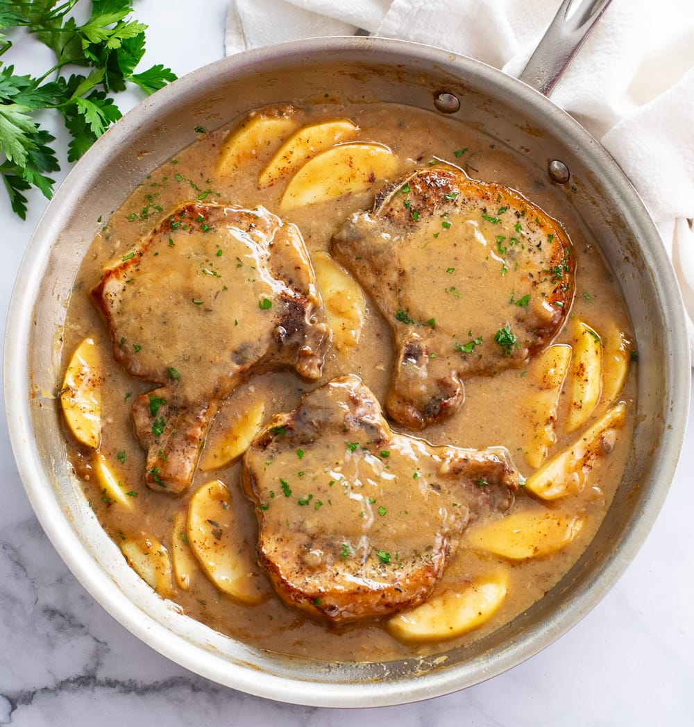 Apple Pork Chops in a skillet with sliced apples and sauce.