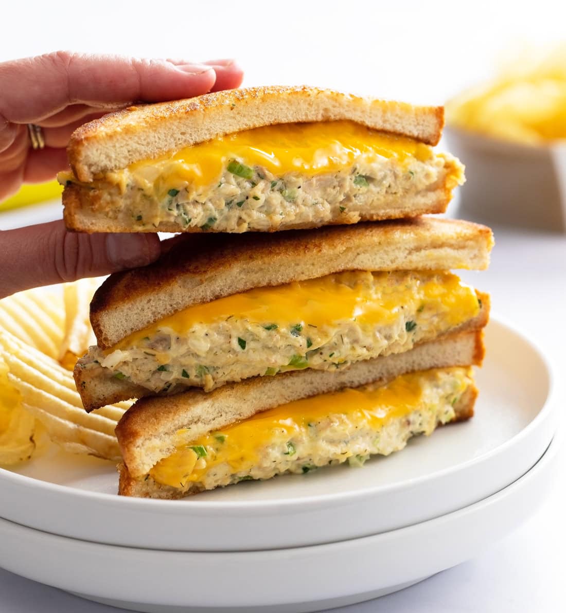 A hand taking the top Tuna Melt from a stack of 3 on a white plate with melted cheese.