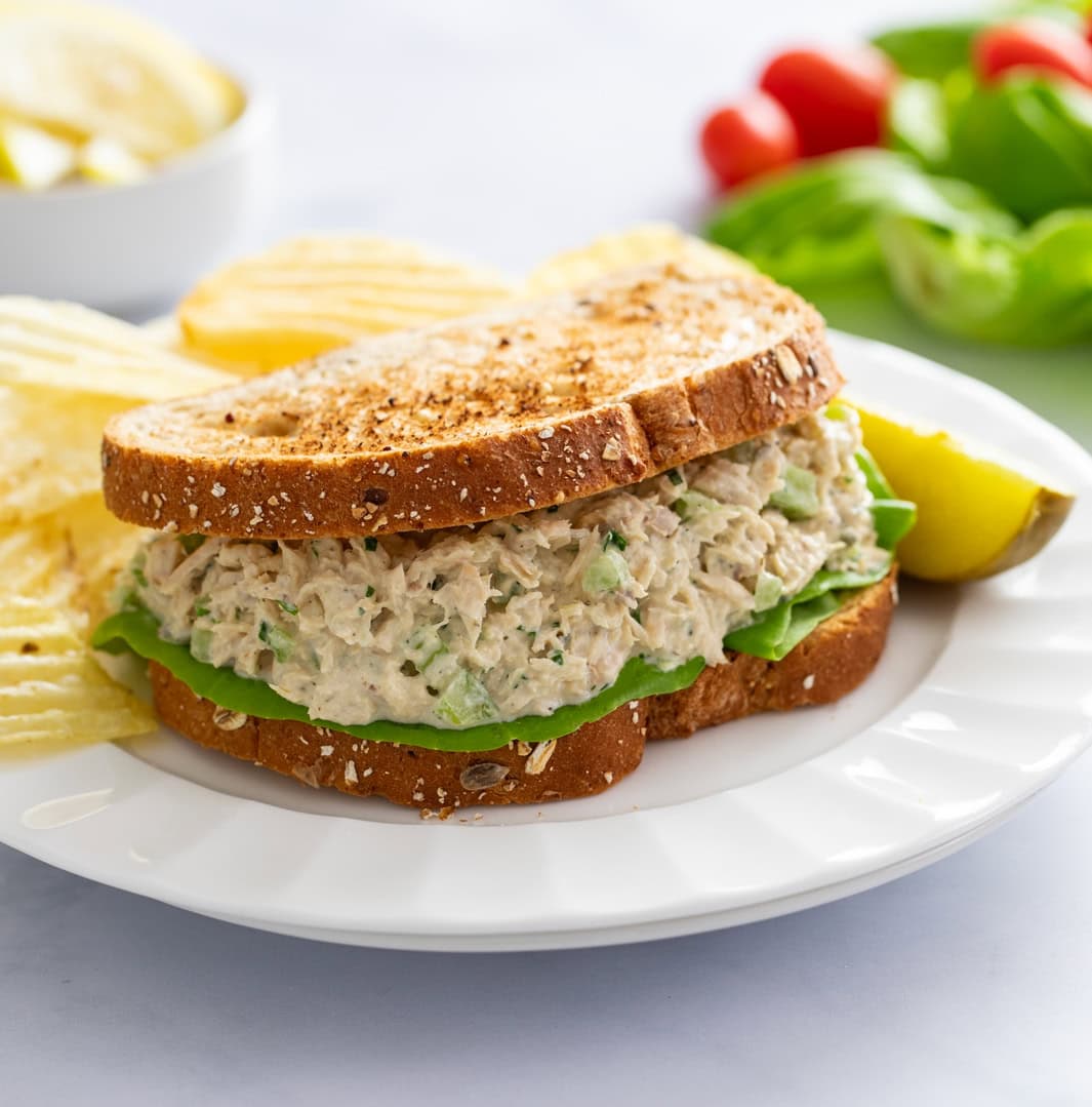 A sandwich filled with Tuna Salad on a white plate with a pickle and chips in the back.