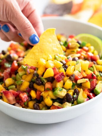 A hand dipping a tortilla chip into a white bowl of Mango Salsa.