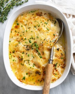 Au Gratin Potatoes in a baking dish topped with chives, thyme, and a serving spoon on the side.
