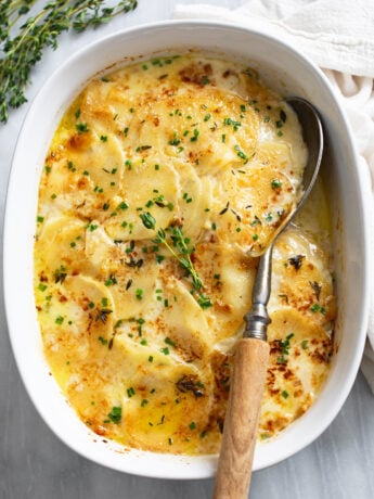 Au Gratin Potatoes in a baking dish topped with chives, thyme, and a serving spoon on the side.