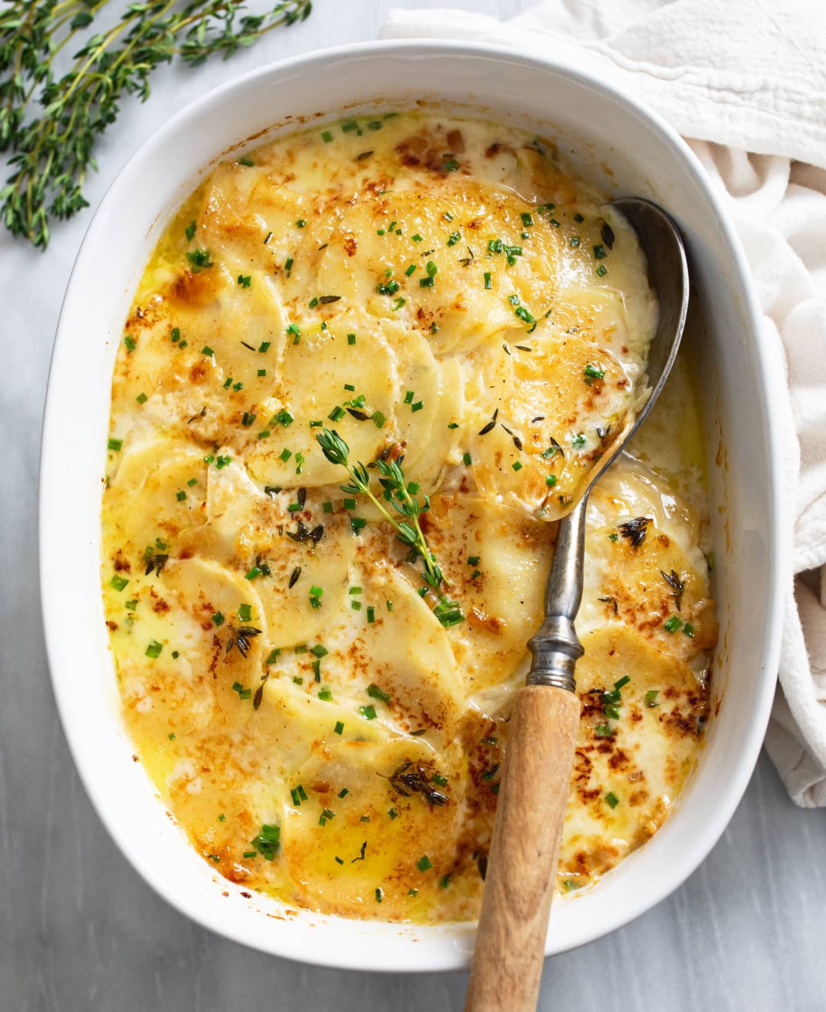 Au Gratin Potatoes in a baking dish topped with chives, thyme, and a serving spoon on the side.