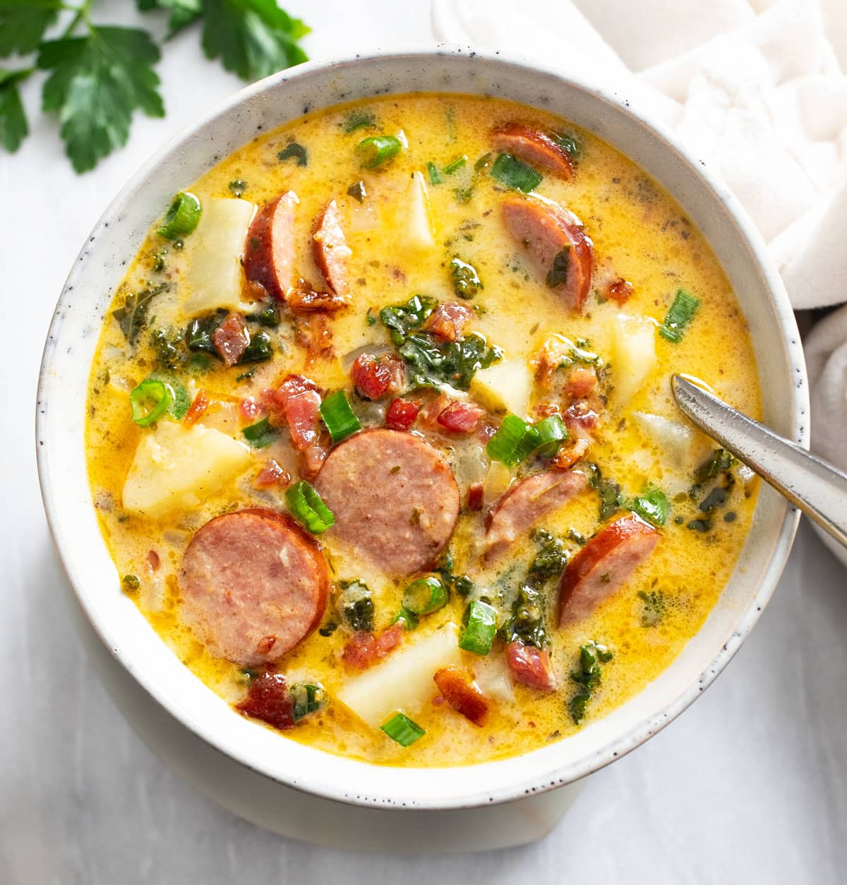 Kielbasa Potato Soup in a soup bowl with a spoon on the side and green onions and bacon on top.