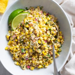 Mexican Street Corn Salad in a white bowl with lime wedges and a spoon on the side.