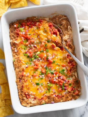 Overhead view of Bean Dip with toppings in a white baking dish with a serving spoon on the side.