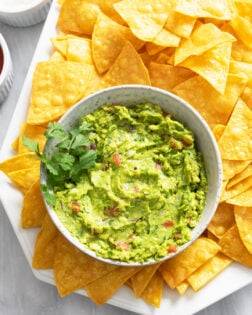 A bowl of Guacamole on a platter with Tortilla Chips and cilantro on the side.