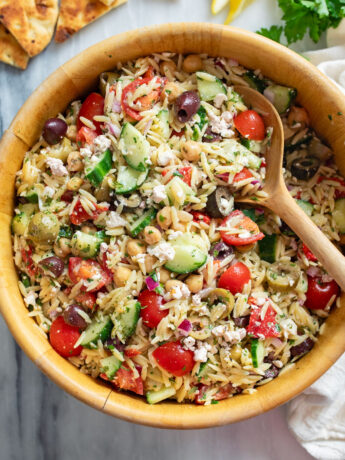 Orzo Salad in a wooden bowl with Mediterranean ingredients and a wooden spoon on the side.