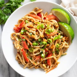 Peanut Butter Noodles in a white bowl with vegetables and lime wedges on the side.