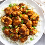 Glossy Orange Chicken over white rice on a white plate with green onions sprinkled on top.