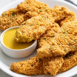A white platter with crispy Baked Chicken Tenders stacked up with one dipped in honey mustard sauce.