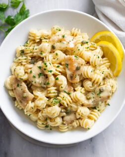 Chicken Piccata Pasta in a white bowl with chopped parsley on top and sliced lemon on the side.