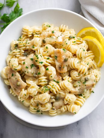 Chicken Piccata Pasta in a white bowl with chopped parsley on top and sliced lemon on the side.
