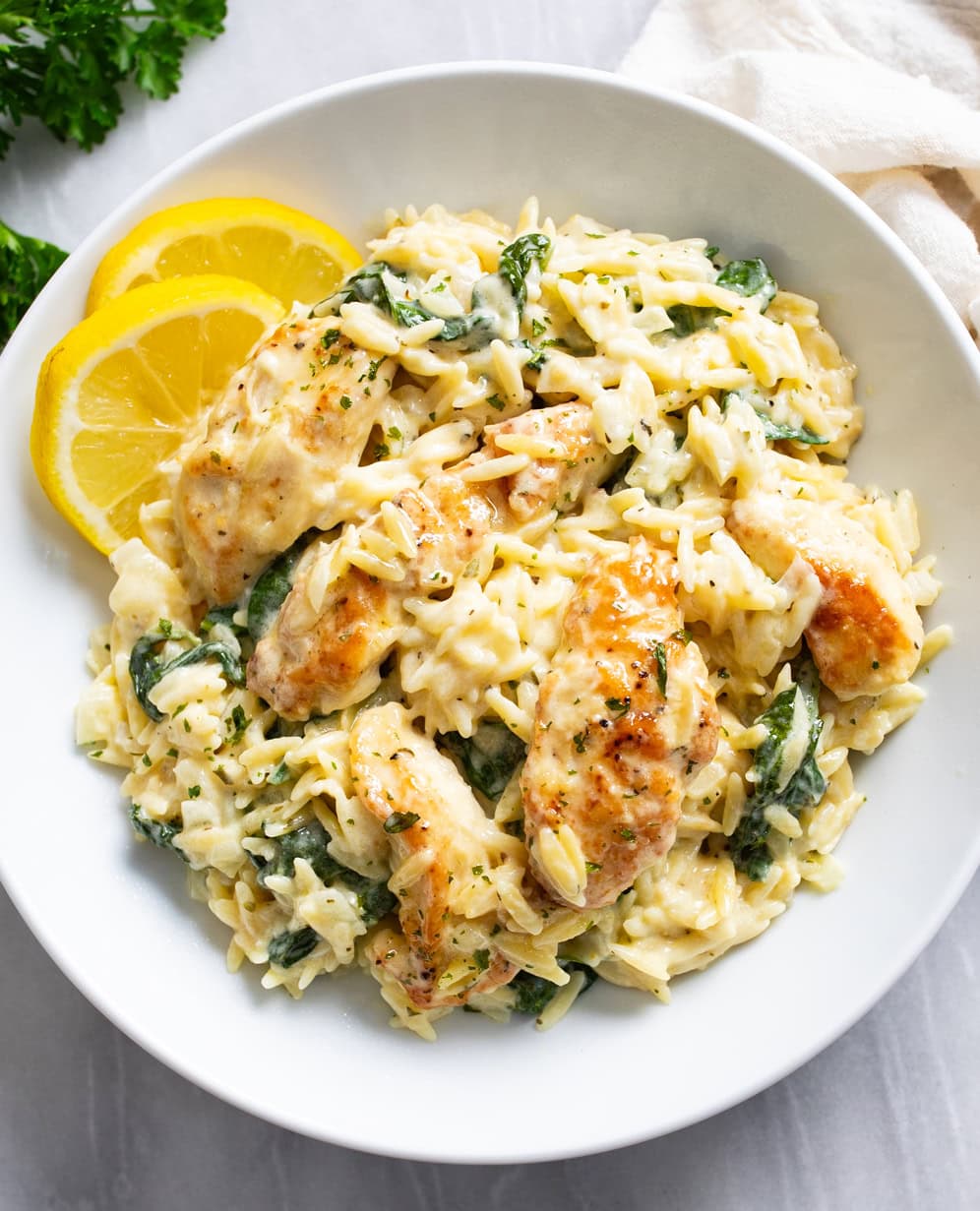 Lemon Chicken Orzo in a white bowl with lemon slices on the side and chicken and spinach in a creamy sauce with orzo.